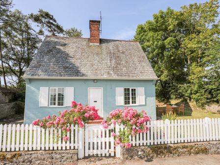 A small house with a pitched roof behind a white picket fence with pink flowers and trees in the garden at Dinam Lodge in Llangaffo