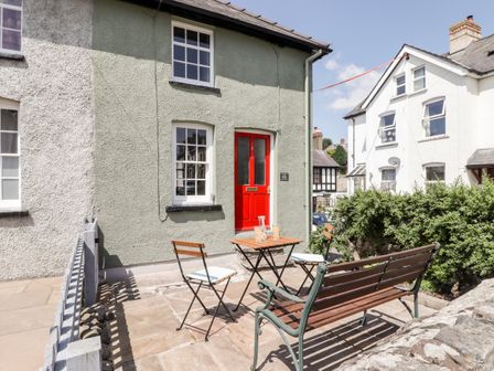 An outdoor patio with two chairs a wooden bench and a small table in front of a green house with a red door at Kooky Cottage in Brecon