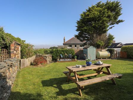 A garden with a wooden picnic table with cups and a teapot a small shed a round table with chairs and a large tree at Hazel Bank Cottage in Penrhyd near Amlwch