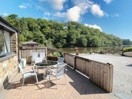 An outdoor patio area with a round glass table and six chairs next to a stone house by a river with trees in the background at Mallard Cottage in Totnes