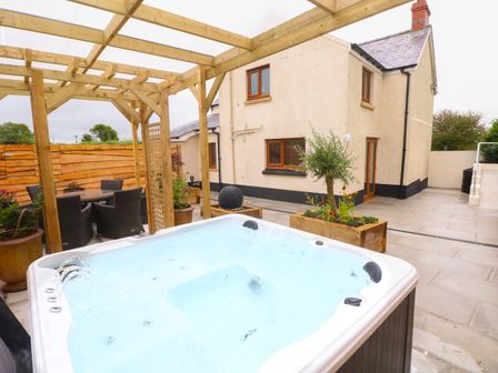An outdoor patio with a hot tub under a wooden pergola and a dining table with chairs next to a beige house at Brynawel in Llangynin near Llanboidy
