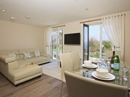 A living room with a beige sofa pillows glass dining table set for four and glass doors opening to a garden at Crowthers in Dartmouth