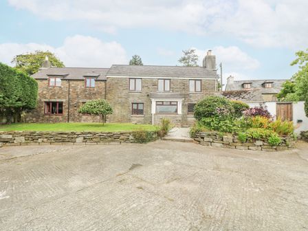 A stone farmhouse with multiple windows a driveway a low stone wall and garden beds with shrubs and flowers at South Boduel Farmhouse in Liskeard