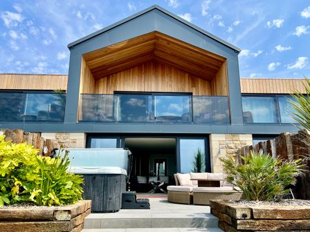 A patio with a hot tub a round outdoor sofa and plants in front of a modern two story house with glass balcony at Sea Scape in Sennen
