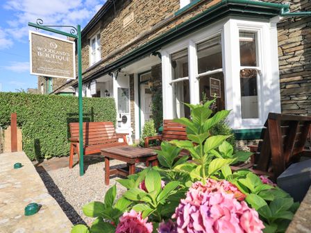 A stone building with wooden benches and a table outside with pink flowers in the foreground at Woodlands Boutique in Windermere