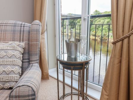 A side table with a bottle in an ice bucket and two glasses next to a window with beige curtains and a plaid armchair at Heron's Landing in Whitby