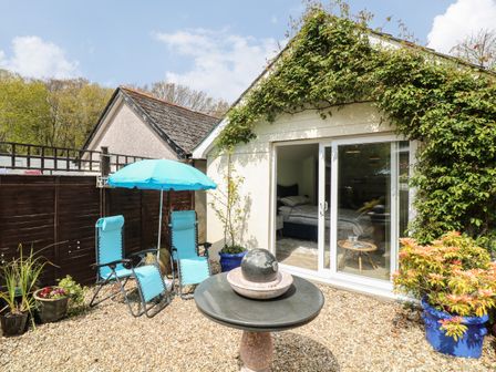 A patio area with two turquoise chairs and umbrella a round stone table with a globe decoration potted plants and a sliding glass door leading to a bedroom at Byghan Thornton in Illogan