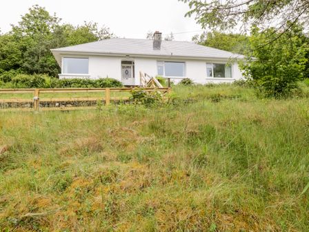A white single-story house with large windows a front door and a wooden fence on a grassy hill at Rainbows End in Clifden County Galway