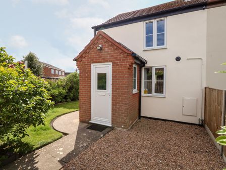 An exterior corner of a house with a red brick porch and white door next to a gravel area and a path leading to a garden at 3 The Cottages in Hemsby