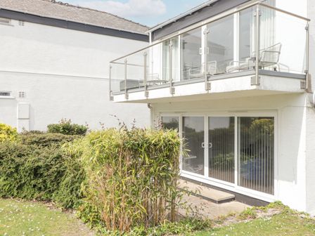 An exterior view of a white building with a glass balcony and sliding glass doors on the ground floor with bushes and grass around at 21 Coedrath Park Saundersfoot
