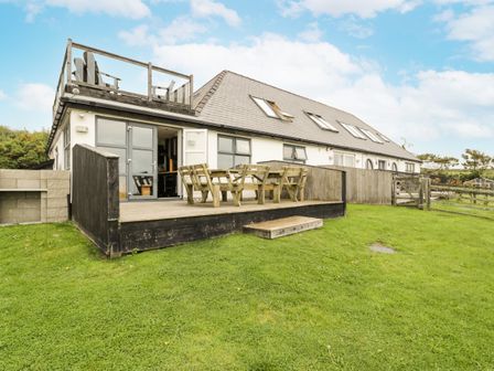 A patio with wooden furniture and a rooftop seating area outside a house with skylights and a grassy lawn at Bwthyn Awel in Church Bay near Cemaes Bay
