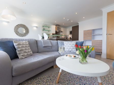 A living room with a grey sofa and patterned cushions a white coffee table with flowers and glasses and a kitchen in the background at The Sanctuary in St Ives