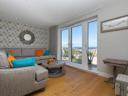 A living room with grey sofas and orange and blue cushions near large windows overlooking a coastal town at The Pearl in St Ives