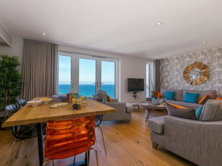 A living and dining area with a wooden table, colorful chairs, grey sofas, a wall mirror, and large windows overlooking the sea at The Oyster in St Ives
