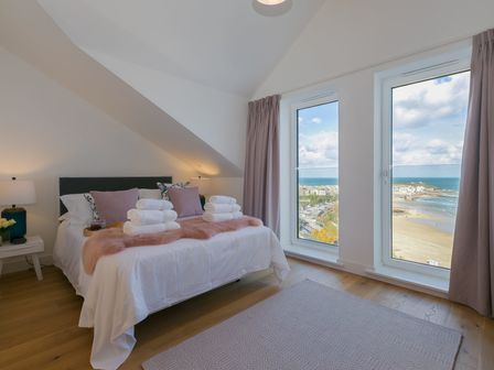 A bedroom with a bed with towels and pillows beside two large windows overlooking the beach at Beach View in St Ives