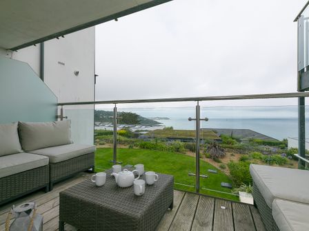A balcony with wicker furniture and a tea set overlooking a garden and the sea at Bright Wings in Carbis Bay