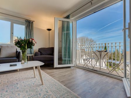 A living room with a gray armchair a white coffee table with flowers and open doors leading to a balcony with a table and chairs and a bottle at Bosbrea in Carbis Bay