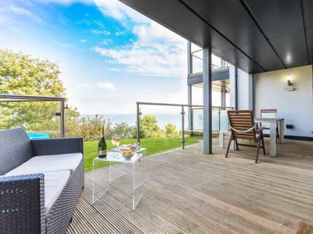 An outdoor balcony with a wicker sofa a table with champagne glasses and wooden chairs overlooking greenery and water at Coast in St Ives