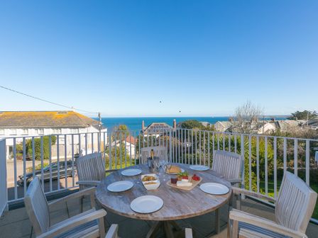 An outdoor balcony with a round wooden table and six chairs overlooking houses and the sea at Cornerways in Carbis Bay