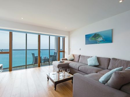 A living room with a large sofa, coffee table with wine glasses, and glass doors opening to a balcony with sea view at Glas Mor in Carbis Bay