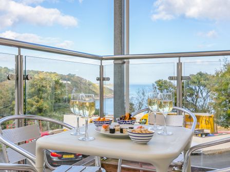 An outdoor balcony with a table set with four glasses of white wine snacks and a view of trees and water at Gwinear in Carbis Bay