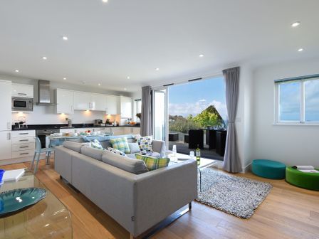An open living room and kitchen with a grey sofa and dining table near a sliding door leading to an outdoor patio at Halcyon Cove in Carbis Bay