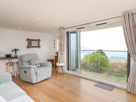 A living room with an armchair a small round table and sliding glass doors opening to a balcony with a sea view at Kernow's Dream in Carbis Bay
