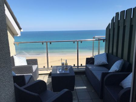 An outdoor balcony with wicker furniture and a glass railing overlooking a sandy beach and ocean at Ocean Cove in Carbis Bay