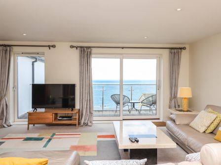 A living room with a TV on a wooden stand a coffee table sofas and chairs with a balcony and ocean view at Ocean View in Carbis Bay