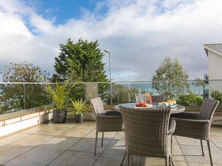An outdoor patio with a round glass table and four wicker chairs with potted plants and a view of trees and the sea at The View in St Ives