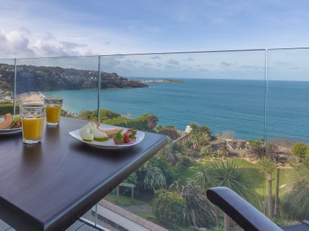 A balcony table with plates of fruit and glasses of juice overlooking the ocean and a garden at Pelagos in Carbis Bay