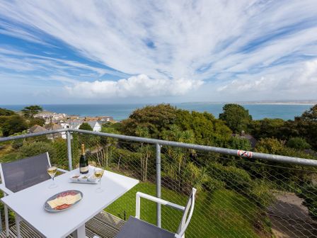 A balcony with a white table and two chairs overlooking the sea and trees at Penthouse View in St Ives