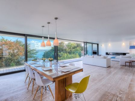 A dining table with white chairs and one yellow chair in a room with large glass windows overlooking trees and water at Salt House in St Ives
