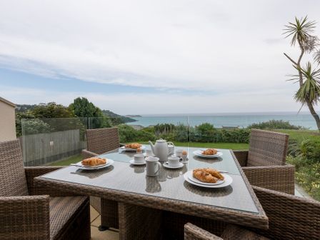 An outdoor dining table with four wicker chairs set with plates of croissants cups and a teapot overlooking a garden and ocean at Seascape in Carbis Bay
