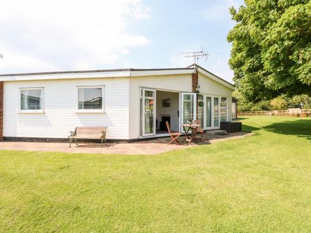 A single-story house with white siding a wooden bench a table with two chairs and a large tree on a grassy lawn at Chalet 150 in Winterton-On-Sea