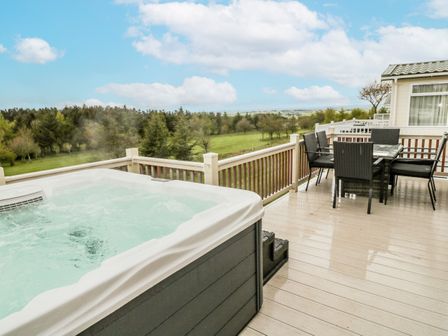 An outdoor deck with a hot tub and a dining table with chairs overlooking a grassy and tree filled landscape at Woodland View Lodge Swarland near Felton