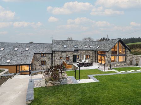A stone house with slate roof and large windows overlooking a patio with outdoor furniture and green lawn at Merddwr in Cerrigydrudion