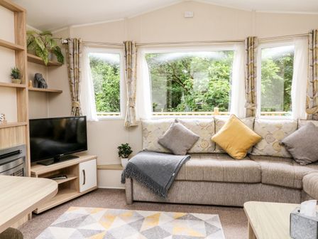 A living room with a beige sofa with cushions beside large windows and a TV on a wooden stand at Ochenta in Combe Martin