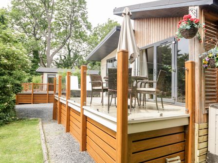 An outdoor wooden deck with glass railing chairs a table and an umbrella next to a house with hanging flower baskets at Robin Lodge in Cartmel