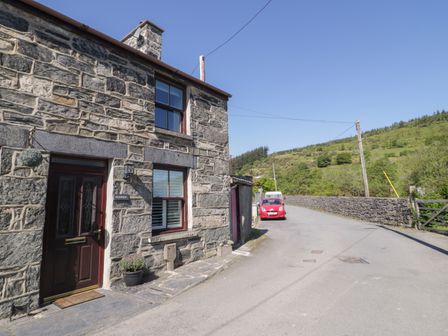 Stone house with a dark wooden door and two windows beside a road with cars and green hills in the background at Dolwen in Penmachno