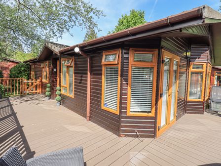 An outdoor wooden deck area with a dark brown wooden cabin with multiple windows and a door at Grizedale Lodge Langdale 18 White Cross Bay near Windermere