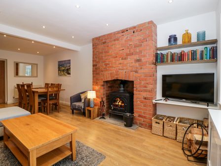 A living room with a wooden dining table and chairs a brick fireplace with a wood stove and a television on a wall shelf at 17 Sands Close in Broadway