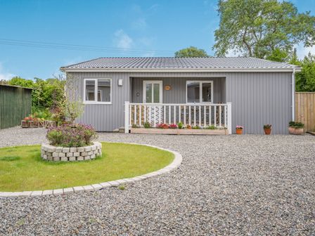 A single story gray house with a small porch and flower beds in front at Caldey Lodge (Bier Wood Lodges) in Manorbier