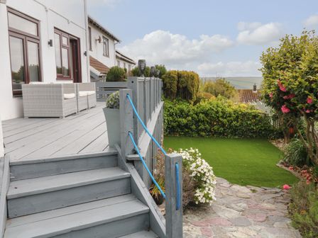 A backyard with a wooden deck, outdoor chairs, stairs, green lawn, plants, and distant hills at 12 Claremont Falls in Polperro