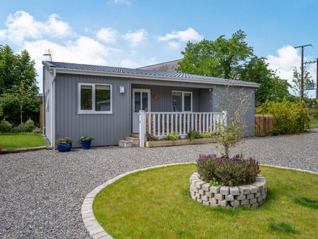 A gray single-story house with a small porch and gravel driveway with a circular planted area in front at Ramsey Lodge Bier Wood Lodges in Manorbier