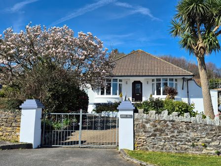 A white house with a tiled roof behind a metal gate and stone wall with trees at Robin Hill in Duporth near St Austell