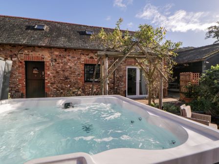 An outdoor hot tub in front of a stone building with wooden doors and windows at The Holt in Harcombe near Sidford