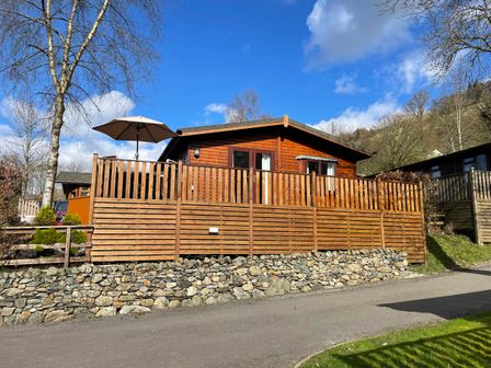 A wooden lodge with a fenced patio and umbrella near a stone wall and trees at Lavender Lodge Applethwaite 4 Limefitt Park near Windermere