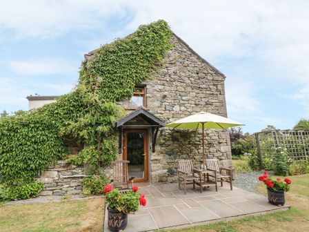 A stone cottage with ivy on the wall wooden chairs and a table with an umbrella on a patio with flower pots at Poppy Cottage in Penruddock