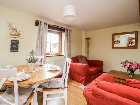 A dining area with a wooden table and white chairs next to a seating area with red sofas and a coffee table with flowers at Lavender Cottage in Penruddock